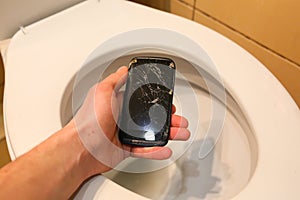 Man is holding a crashed black smartphone in hand over the toilet bowl. Broken lcd touch screen in the bathroom. Information