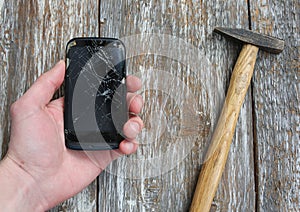 Man is holding a crashed black smartphone in hand on the wooden background. Broken lcd touch screen with a hammer. Information