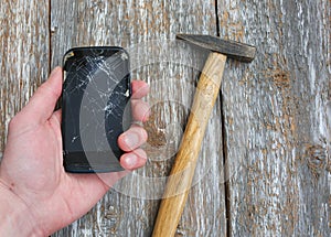 Man is holding a crashed black smartphone in hand on the wooden background. Broken lcd touch screen with a hammer. Information