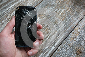 Man is holding a crashed black smartphone in hand on the wooden background. Broken lcd touch screen. Information technology photo