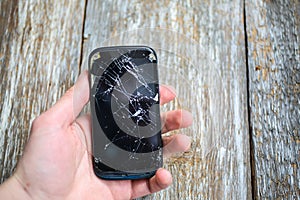 Man is holding a crashed black smartphone in hand on the wooden background. Broken lcd touch screen. Information technology photo