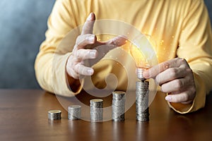 Man holding a glowing abstract light bulb with pile money cions on a wooden table, business problem solving ideas and creative