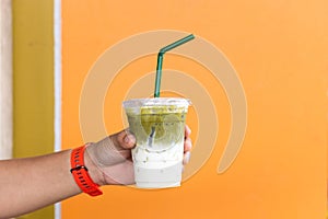 A man holding green tea with milk and iced cup in hand with orange color wall