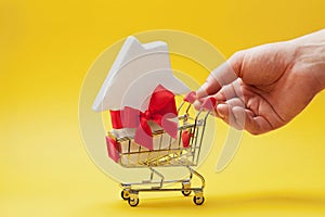 Man holding hand shopping cart with house decorated ribbon on yellow background. Buying a new home, gift or sale of real estate.