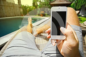 Man is holding his smartphone with a blank screen in his hand on the background of the pool and his feet in a deckchair.