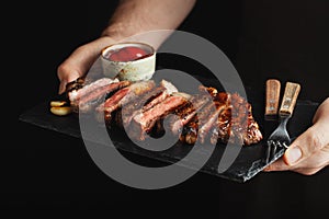 Man holding juicy grilled beef steak with spices and red sauce on a stone cutting board on a black background