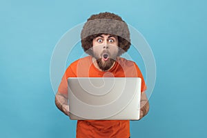 Man holding laptop and looking at camera with open mouth and big eyes.