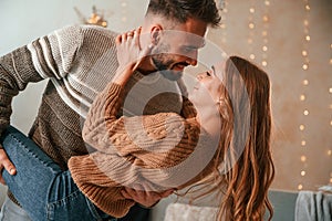 Man is holding leg of the woman while standing. Lovely young couple are celebrating New Year at home