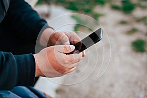 Man holding mobile phone outdoor. Traveler with black hoodie using smarphone sitting outside. photo from aside