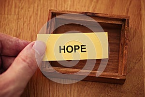 A man holding a piece of yellow paper with the word Hope on it in his hand over a small empty wooden box.