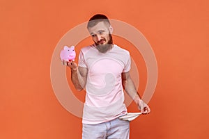 Man holding piggybank in hand and showing his empty pockets, being frustrated and upset.