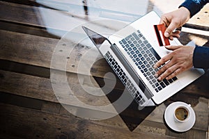 Man holding plastic credit card in hand and typing laptop keyboard while sitting at the wooden table.Reflections on