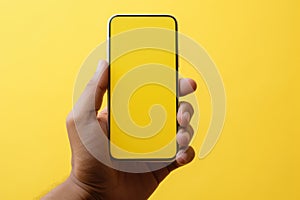 Man holding smartphone with blank screen on yellow background. Closeup of hand with cell phone. Mockup phone screen