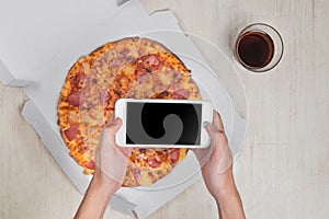 Man holding smartphone and tasty pizza with ingredients on wooden table.