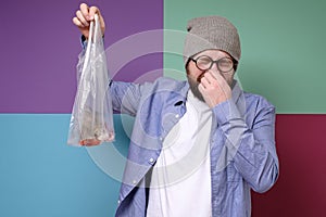 Man is holding a smelly fish in a transparent package, pinches fingers nose because of bad smell and closes his eyes.