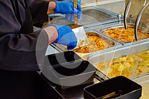 Man holding a spatula serving food on an empty plastic food container in a cafeteria