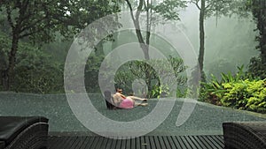 Man holding and spinning young woman in infiniti pool overlooking tropical misty jungle view during rain. Beautiful