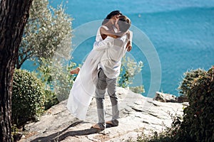 Man holding woman in his arms wrapped around her waist and she is holding on to him, standing near sea in Greece.