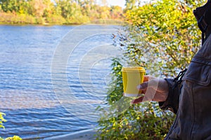 Man holding yellow cup on river background. Coffee cup in nand on autumn landscape background. Travel concept. Vacations at nature