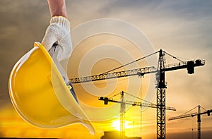 Man holding the yellow safety helmet at construction site with crane background silhouette