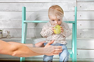 Man holds baby boy who eat big green apple. He both are in jeans and white hoodie. Infant sits on the steps indoor. White wooden