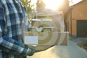 Man holds blank box, coffee cups and package outdoor. Delivery