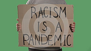 A man holds in front of him a poster from a cardboard box with the words RACISM IS A PANDEMIC. Isolated a green screen