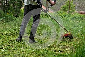 A man holds a grass trimmer and cuts tall grass in his yard on a spring morning. Close up of the farmer in boots while working in