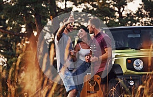 Man holds guitar. Group of cheerful friends have nice weekend at sunny day near theirs green car outdoors