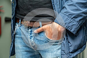 A man holds his hand in a jeans pocket with a leather belt. Blue jacket, black T-shirt. Horizontal orientation.