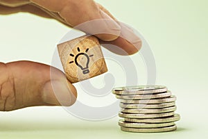 A man holds in his hand a wooden cube with a picture of a light bulb near a stack of coins