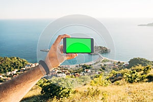 Man holds horizontally in his hand a smartphone with a blank screen against the background of Sveti Stefan island