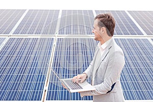 A man holds an laptop in his hands and looks in the distance, standing next to solar panels. Outdoors