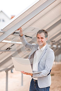 A man holds an laptop, and holds his hand behind the solar panel and smiles.