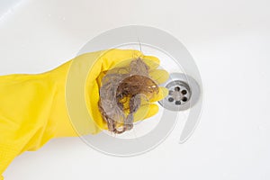 A man holds a lot of hair in rubber yellow gloves against the background of a sink. The problem of clogged drains in the photo