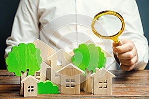 A man holds a magnifying glass over wooden houses and trees. Real estate valuation and selection of a place for the construction