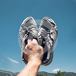 Man Holds Old Running Sneakers In Hand On Background Of Bue Sky And Mountains