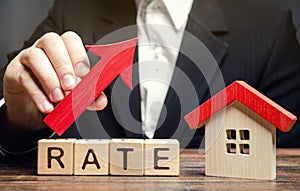 A man holds a red arrow up above the word Rate and a wooden house. The concept of raising interest rates on mortgages. The