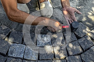 A man holds a red-black rubber mallet in his hand and knocks it on the surface of gray granite stones on the sidewalk, top view.