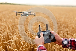 Man holds remote controller with his hands while copter is flying on background. Drone hovers behind the pilot in wheat