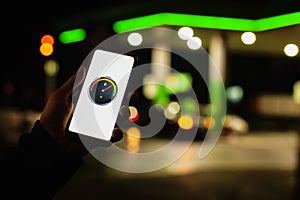 A man holds a smartphone with a digital fuel meter on the screen against the background of a night gas station for a car