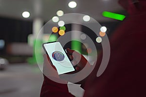 A man holds a smartphone with a digital fuel meter on the screen against the background of a night gas station for a car