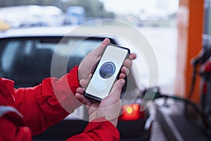 Man holds a smartphone with a digital fuel meter on the screen in the background of a gas station and a car