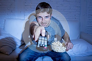 Man at home lying on couch at living room watching tv eating popcorn bowl using remote control