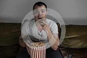 A man at home sitting on the couch watching TV and eating popcorn