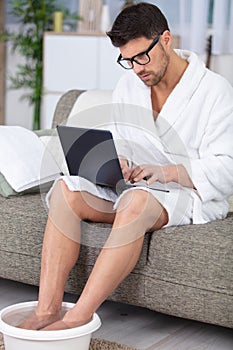 man at home soaking feet and using laptop