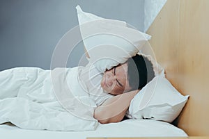 A man at home tries to fall asleep, noisy neighbors interfere with sleep, an Asian closes his ears with pillows, tired after work