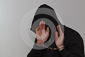 A man with a hood on his head with his hands cuffed hides his face against a gray background.