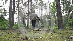 A man in a hood and a vest rolls a bicycle through the forest. Cycling and travel concept, slow mo