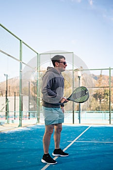 A man in a hoodie and shorts smiles while holding a padel tennis racket on a sunny outdoor court with mountains in the